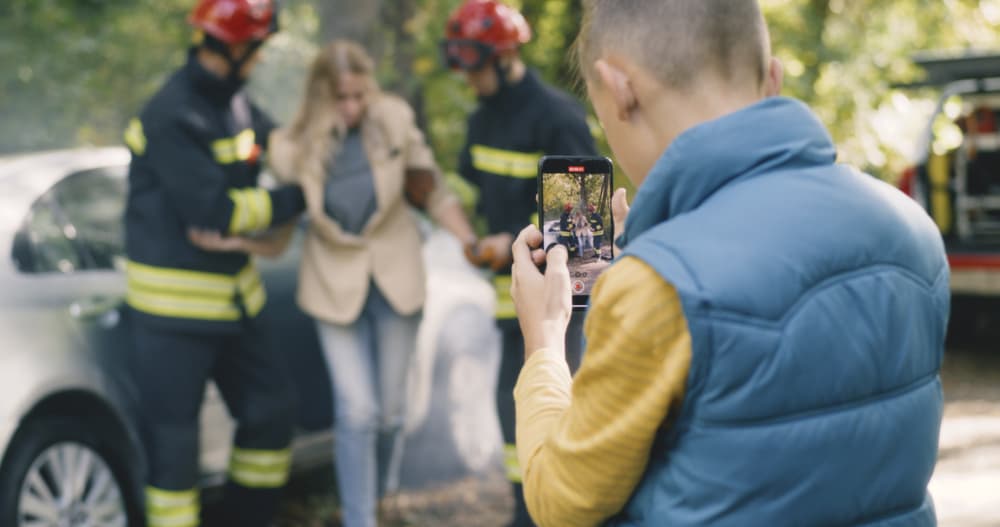 Boy in waistcoat using smartphone to shoot firefighters helping blond woman to get out wrecked car after car crash